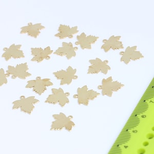 May include: A collection of 18 gold-colored metal maple leaf charms. The charms are scattered on a white surface with a green ruler in the background.