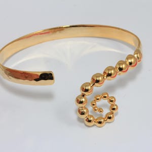 May include: A gold-colored cuff bracelet with a hammered texture on the band. The bracelet features a spiral design made of small, round beads. The open-ended design allows for easy wear. The bracelet is on a white surface.