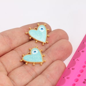 Könnte beinhalten: Zwei goldfarbene herzförmige Anhänger mit einer hellblauen Emaille-Oberfläche und einem blauen bösen Blick-Anhänger in der Mitte jedes Herzens.