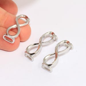 Könnte beinhalten: Drei silberne Charms in Form eines Unendlichkeitszeichens, zwei liegen flach und einer wird in der Hand gehalten. Die Charms sind glatt und glänzend.
