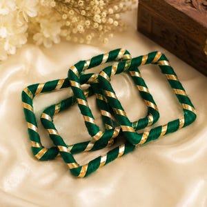 May include: Four square bangles with a green and gold spiral design. The bangles are made of a soft, textured material and are arranged on a cream-colored satin surface. A wooden box is in the background.