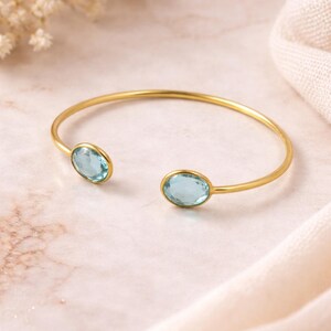 Può includere: Un bracciale aperto color oro con due pietre preziose ovali azzurro chiaro. Il bracciale è esposto su una superficie di marmo rosa chiaro con un morbido tessuto bianco sullo sfondo. Sono visibili delicati fiori bianchi.