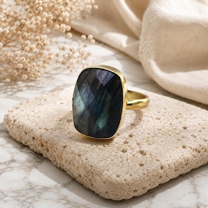 May include: A gold ring featuring a large, rectangular, faceted labradorite gemstone with a dark grey surface that displays iridescent blue and green flashes. The stone is set in a smooth gold metal bezel with a simple, polished gold band.