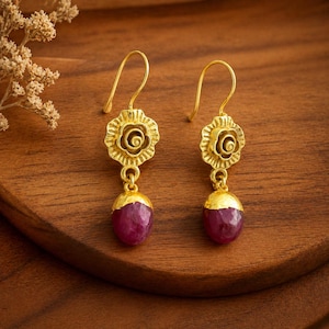 May include: A pair of gold-colored earrings with a floral design at the top and a teardrop-shaped, deep red gemstone at the bottom. The earrings are displayed on a wooden surface.