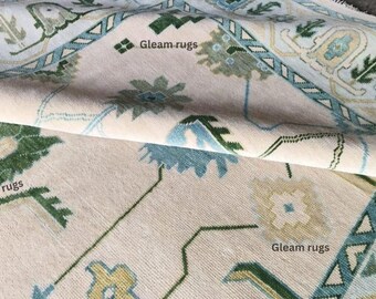 May include: Close-up of a beige rug with a floral and geometric pattern in shades of green, blue, and yellow. The rug has a rolled edge, revealing the underside. The text "Gleam rugs" is visible.