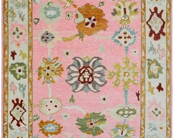 May include: A pink rug with a floral pattern. The rug has a light blue border with a gold trim. The rug is made of wool and is hand-knotted.
