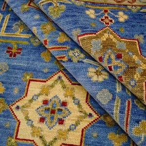 May include: A folded blue rug with intricate geometric patterns. The rug features a central design with a cream-colored starburst shape, surrounded by red, yellow, and green accents. The overall design is symmetrical and detailed.