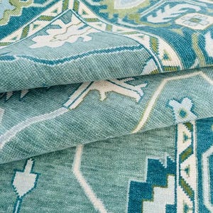 May include: A teal blue rug with a white and green geometric pattern. The rug is folded in half, showing the intricate details of the design.