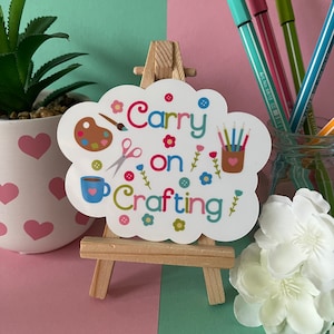 May include: White sticker with a cloud shape and the text "Carry on Crafting" in colourful letters. The sticker has various crafting supplies like paintbrushes, scissors, buttons, and a cup of coffee.