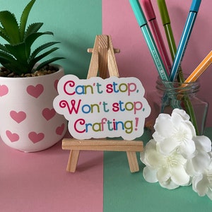 May include: A sticker on a wooden easel reads "Can't stop, Won't stop, Crafting!" The sticker has a colorful, whimsical font. The easel is on a pink and green surface.