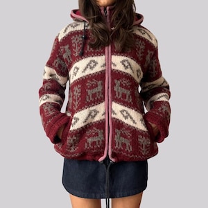 Könnte beinhalten: Ein weinroter und cremefarbener Kapuzenpullover mit Reißverschluss und einem Muster aus Hirschen und geometrischen Formen. Der Pullover hat einen rosa Reißverschluss und eine Kapuze mit Kordelzug. Die Ärmel haben Taschen und wird über einem Jeansrock getragen.