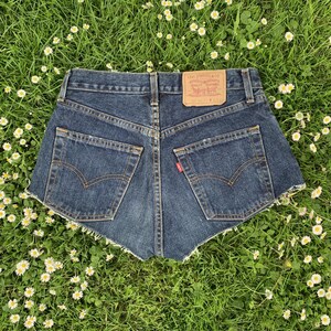 May include: Dark blue denim shorts with frayed edges. The shorts have a back pocket with a red Levi's tab and a brown leather patch with the Levi's logo and text that reads "Levi Strauss & Co. San Francisco, Calif. Original Quality Clothing. WPL 423."