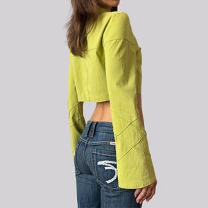 Könnte beinhalten: Ein kurz geschnittenes, langärmeliges chartreusefarbenes Oberteil mit gesteppten Nahtdetails an den Ärmeln. Das Oberteil wird mit einer blauen Jeans kombiniert. Die Jeans hat ein weißes Stickmuster auf der Gesäßtasche.