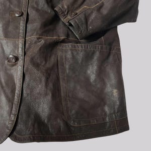 May include: A brown leather jacket with a button-up front and a pocket. The jacket has visible stitching and a worn appearance. The sleeve is partially visible, showing button details. The jacket is a medium brown color.