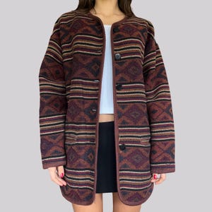 Könnte beinhalten: Lange, geknöpfte Jacke mit geometrischem Muster in Braun-, Burgunder-, Schwarz- und Beigetönen. Die Jacke hat einen runden Kragen und zwei Taschen. Die Jacke ist aus einem dicken, gewebten Material gefertigt.