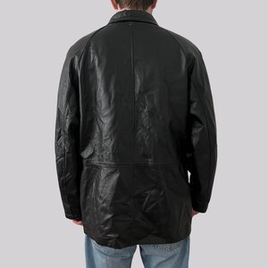 May include: A black leather jacket with a collared neckline and long sleeves. The jacket has a button closure and a tailored fit. The back of the jacket is visible, showing the seams and the texture of the leather. The jacket is worn over blue jeans.