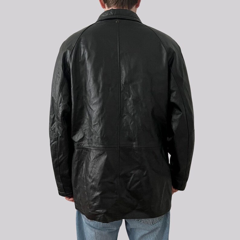 May include: A black leather jacket with a collared neckline and long sleeves. The jacket has a button closure and a tailored fit. The back of the jacket is visible, showing the seams and the texture of the leather. The jacket is worn over blue jeans.