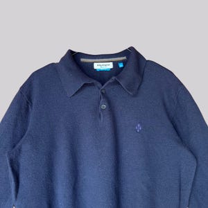 K&ouml;nnte beinhalten: Marineblaues Langarm-Poloshirt mit Kragen und Knopfleiste. Das Hemd hat ein kleines rautenf&ouml;rmiges Emblem auf der Brust und ein Markenetikett am Kragen. Der Stoff scheint ein weicher, strukturierter Strick zu sein.