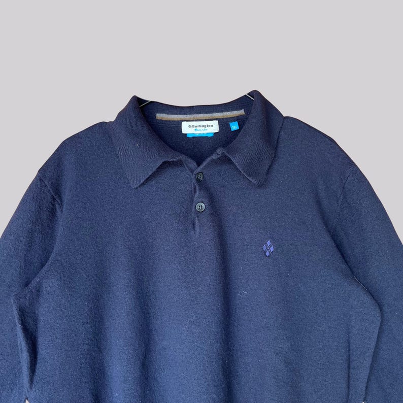K&ouml;nnte beinhalten: Marineblaues Langarm-Poloshirt mit Kragen und Knopfleiste. Das Hemd hat ein kleines rautenf&ouml;rmiges Emblem auf der Brust und ein Markenetikett am Kragen. Der Stoff scheint ein weicher, strukturierter Strick zu sein.