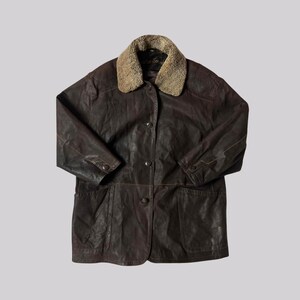May include: A brown leather jacket with a tan shearling collar. The jacket has a button-up front, two front pockets, and buttoned cuffs. The jacket appears to be vintage.