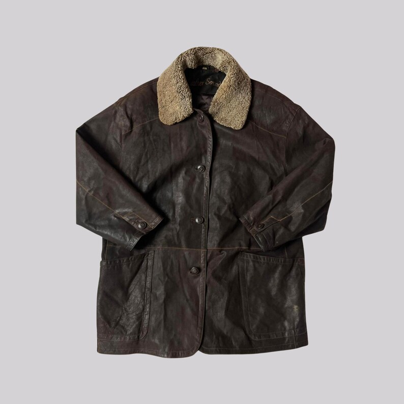 May include: A brown leather jacket with a tan shearling collar. The jacket has a button-up front, two front pockets, and buttoned cuffs. The jacket appears to be vintage.