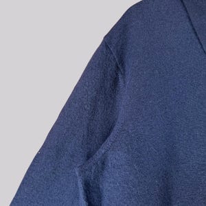 K&ouml;nnte beinhalten: Nahaufnahme eines marineblauen Pullovers. Der Stoff hat eine strukturierte Optik, und der Halsausschnitt ist sichtbar. Der Pullover scheint aus einem weichen Material zu sein, das f&uuml;r k&uuml;hlere Temperaturen geeignet ist. Das Bild ist gut beleuchtet und hebt die Details des Strickwerks hervor.
