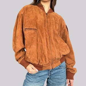 May include: A brown suede bomber jacket with a zip-up front, a small chest pocket, and two side pockets. The jacket has brown ribbed cuffs and a brown ribbed collar. The jacket is a vintage style.