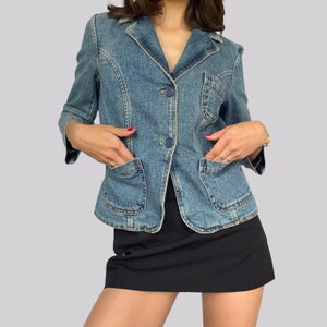 May include: A blue denim jacket with a button closure and two front pockets. The jacket is cropped and has a collar.