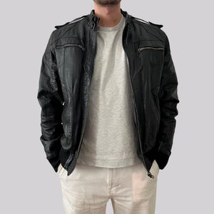 May include: Black leather jacket with a zip-up front, multiple pockets, and a ribbed hem. The jacket has a classic biker style with a stand-up collar and shoulder accents. The model is wearing a gray t-shirt and light-colored pants.
