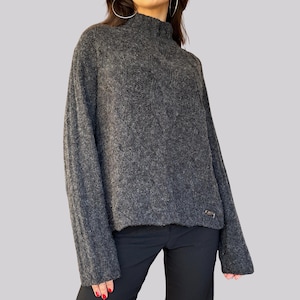 Puede incluir: Un suéter de punto trenzado gris oscuro con cuello alto. El suéter de manga larga tiene un patrón texturizado y una pequeña etiqueta metálica cerca del dobladillo. La modelo lleva pantalones negros.