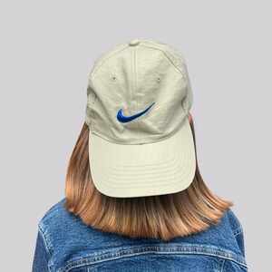 May include: A light beige baseball cap featuring a vibrant blue embroidered Nike swoosh logo on the front centre. The cap has a structured six-panel design with a curved brim and stitched eyelets for ventilation, providing a classic athletic look.
