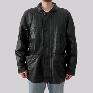 May include: A black leather jacket with a button-up front, two front pockets, and a collared neckline. The jacket has a classic design and appears to be vintage. The jacket is worn over a white shirt.