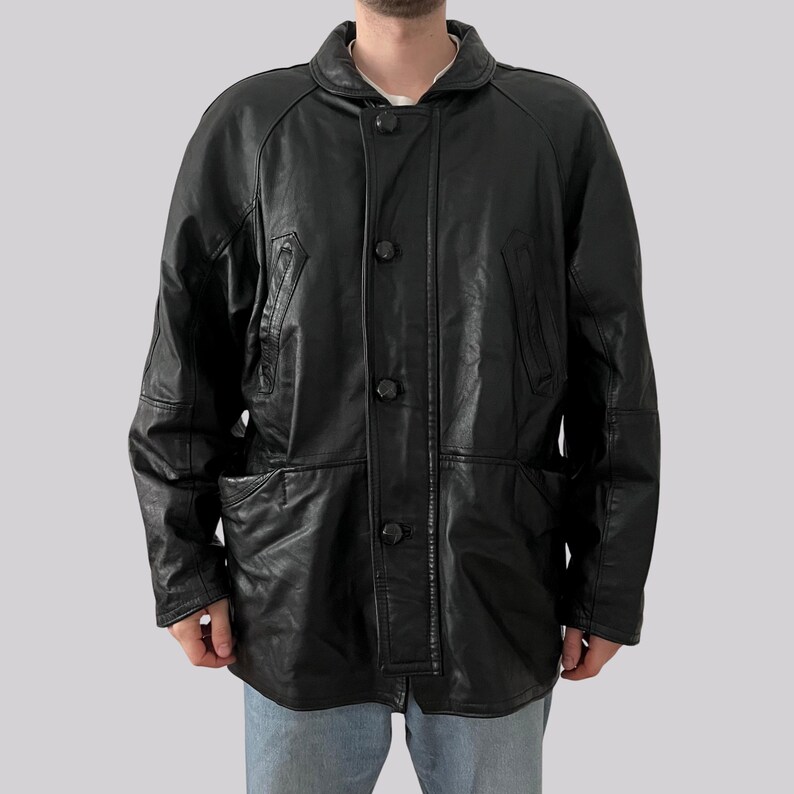 May include: A black leather jacket with a button-up front, two front pockets, and a collared neckline. The jacket has a classic design and appears to be vintage. The jacket is worn over a white shirt.