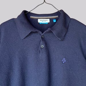K&ouml;nnte beinhalten: Ein marineblaues Poloshirt mit Kragen und drei Kn&ouml;pfen. Das Hemd hat ein kleines gesticktes Emblem unten rechts und ein Etikett mit der Aufschrift "Burlington". Das Hemd h&auml;ngt an einem schwarzen Kleiderb&uuml;gel.