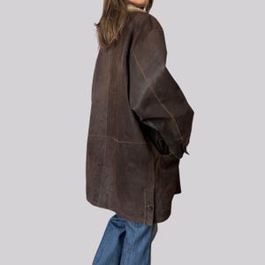 May include: A dark brown leather coat with a button closure and two front pockets. The coat has a shearling collar and is worn with blue jeans. The coat appears to be vintage.