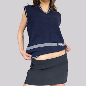 May include: Navy blue sleeveless sweater vest with a V-neck and white trim. The vest has a horizontal white stripe design across the chest with the word "Puma" printed on the right side. The model is wearing a black skirt.