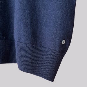 K&ouml;nnte beinhalten: Marineblauer Strickpullover mit gerippten B&uuml;ndchen und einem kleinen, dekorativen Knopf am Saum. Die Nahaufnahme zeigt die Struktur des Stoffes und das Design des Knopfes. Ein klassisches, vielseitiges Kleidungsst&uuml;ck.