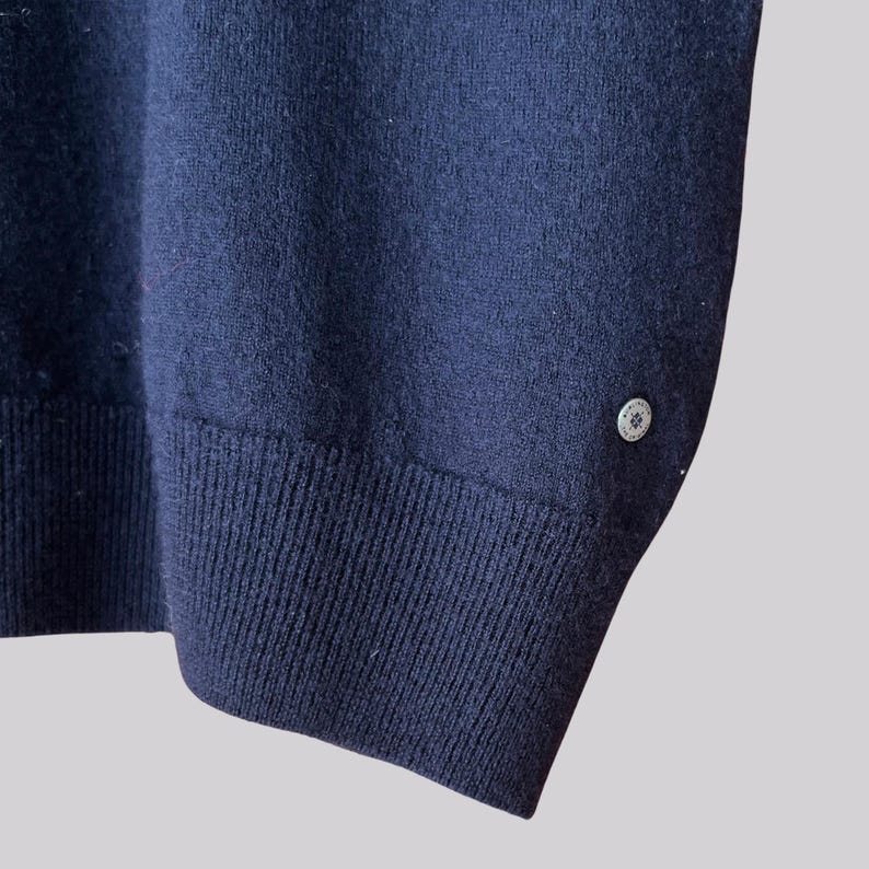 K&ouml;nnte beinhalten: Marineblauer Strickpullover mit gerippten B&uuml;ndchen und einem kleinen, dekorativen Knopf am Saum. Die Nahaufnahme zeigt die Struktur des Stoffes und das Design des Knopfes. Ein klassisches, vielseitiges Kleidungsst&uuml;ck.