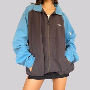 Könnte beinhalten: Eine schwarz-blaue Farbblockjacke mit Reißverschluss und dem Schriftzug "FILA" in Weiß auf der Vorderseite.