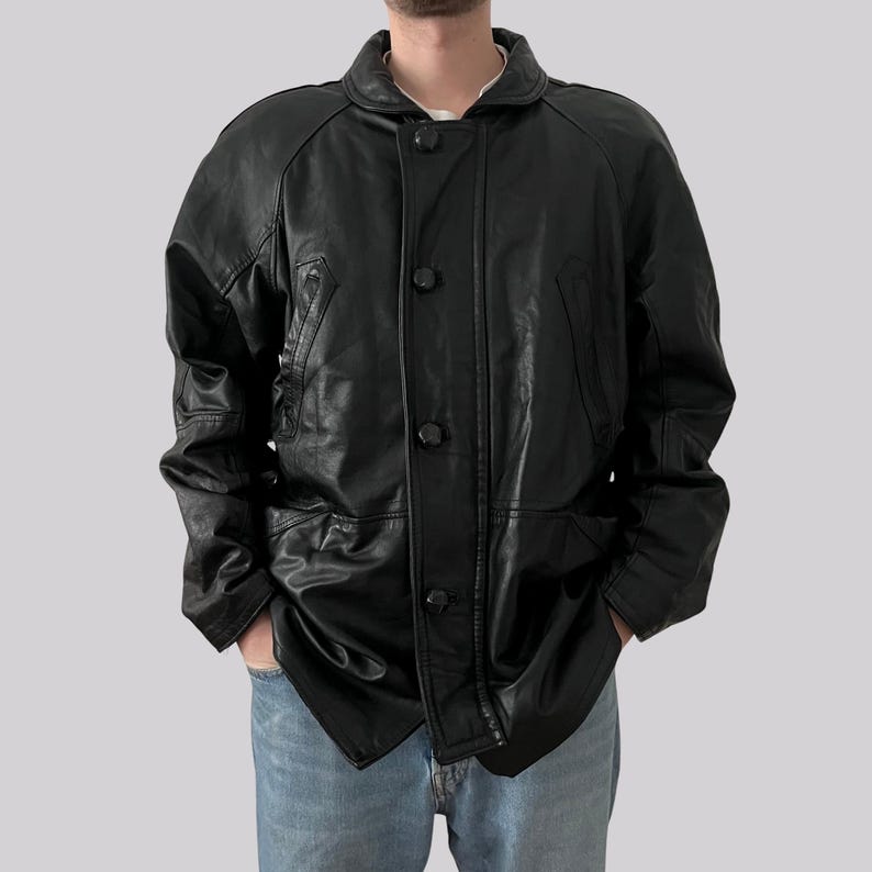May include: A black leather jacket with a button-up front, two front pockets, and a collared neckline. The jacket has a classic design and appears to be vintage. The model is wearing blue jeans.