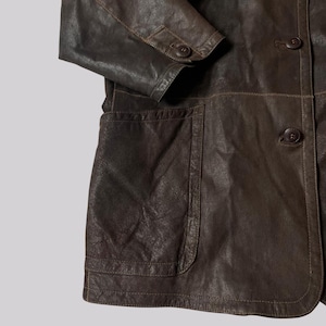May include: A close-up of a brown leather jacket with a buttoned front and two front pockets. The jacket has a classic design with visible stitching and a textured surface. The sleeve is folded at the elbow.