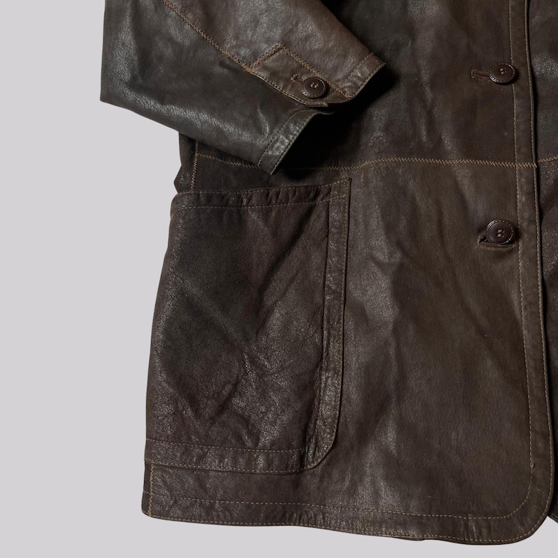 May include: A close-up of a brown leather jacket with a buttoned front and two front pockets. The jacket has a classic design with visible stitching and a textured surface. The sleeve is folded at the elbow.