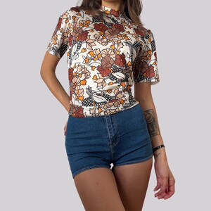 Może przedstawiać: Biała koszula z krótkim rękawem z brązowym i pomarańczowym nadrukiem kwiatowym. Koszula ma wysoki kołnierz i dopasowany krój.