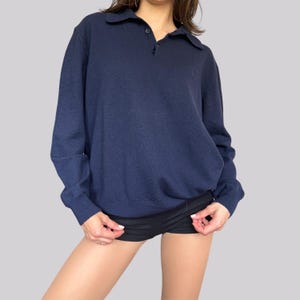 K&ouml;nnte beinhalten: Ein marineblaues Polohemd mit langen &Auml;rmeln, Kragen und Knopfleiste. Das Hemd hat ein kleines Logo auf der Brust. Das Model tr&auml;gt schwarze Shorts.