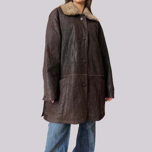May include: A dark brown leather coat with a shearling collar. The coat has a button-up front, two front pockets, and a knee-length hem. The coat is worn over blue jeans.