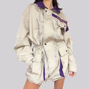 Könnte beinhalten: Eine beige Jacke mit einem violetten und goldenen Besatz. Die Jacke hat einen Kordelzug in der Taille und mehrere Taschen. Der Text "BENSON" ist auf der Jacke sichtbar.