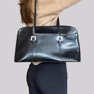 May include: A black rectangular shoulder bag with a smooth, glossy finish. The bag features two long shoulder straps and two decorative silver-toned metal buckles on the front panel, positioned symmetrically above vertical stitched seams.