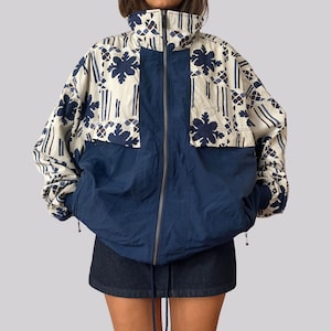 Op de afbeelding: Een jack met ritssluiting, voorzien van een effen marineblauw onderste gedeelte en een crèmekleurig bovenste gedeelte met een donkerblauw bloemen- en geometrisch patroon. Het jack heeft een hoge kraag, lange mouwen met geribbelde boorden en een rits.