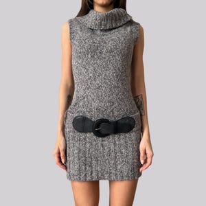 May include: A sleeveless grey knitted dress with a roll neck collar and a black leather belt. The dress has a textured appearance and a fitted silhouette. The belt features a large, round buckle. The dress is a mini-length.