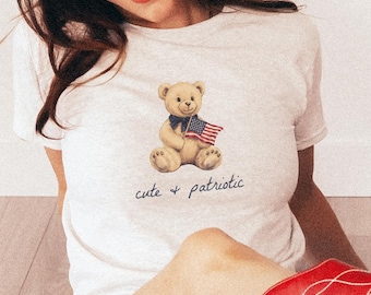 May include: A light grey t-shirt with a teddy bear wearing an American flag, paired with red cowboy boots and red and white checkered shorts. The text "cute + patriotic" is printed on the shirt.